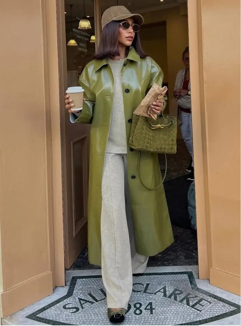 Woman in a green trench coat and beige hat holding a coffee cup and handbag, standing on a street.