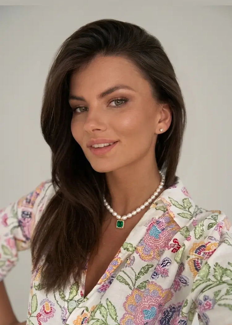 A woman with long dark brown hair is shown from the chest up against a plain light gray background. She is wearing a white blouse with a colorful floral and butterfly print, a pearl necklace with a green rectangular pendant, and small gold hoop earrings.