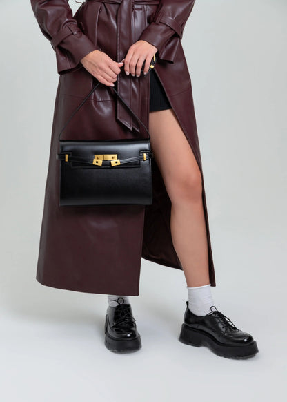 Person wearing a long brown leather coat, black shoes, and holding a black handbag on a white background
