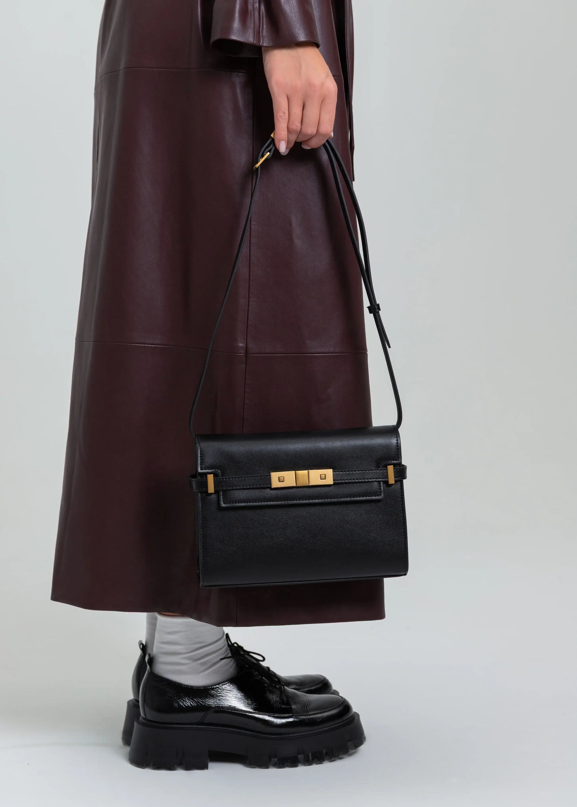 Person wearing a long dark coat holding a black handbag on a light gray background