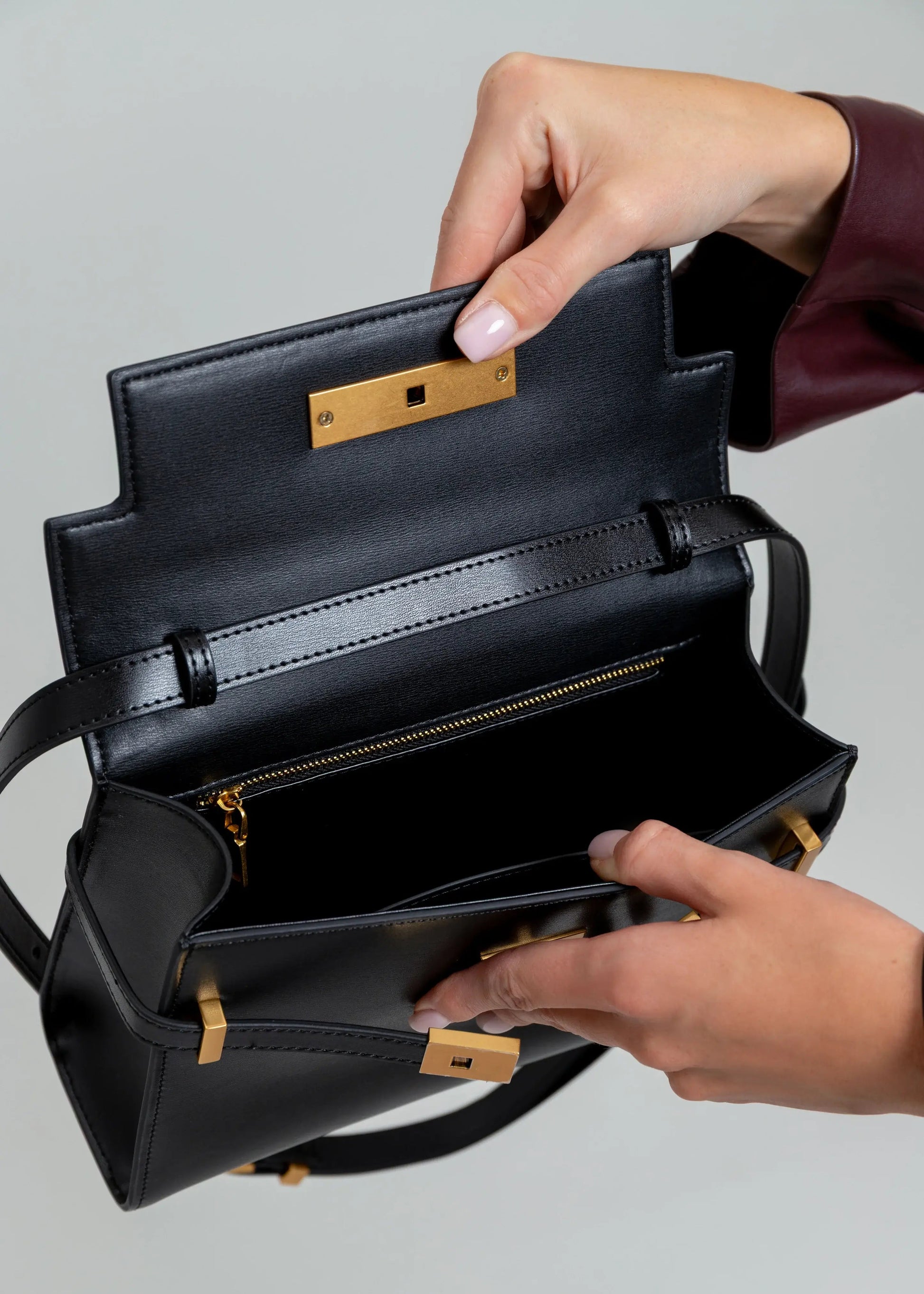 Black handbag with gold accents held open by a person's hands on a gray background
