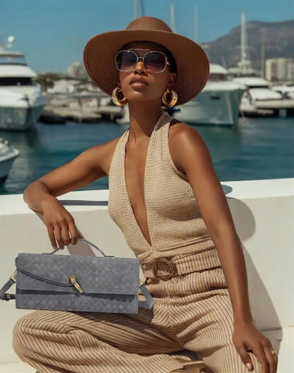 Woman in a beige outfit with a wide-brimmed hat and sunglasses, sitting on a boat with boats and mountains in the background.