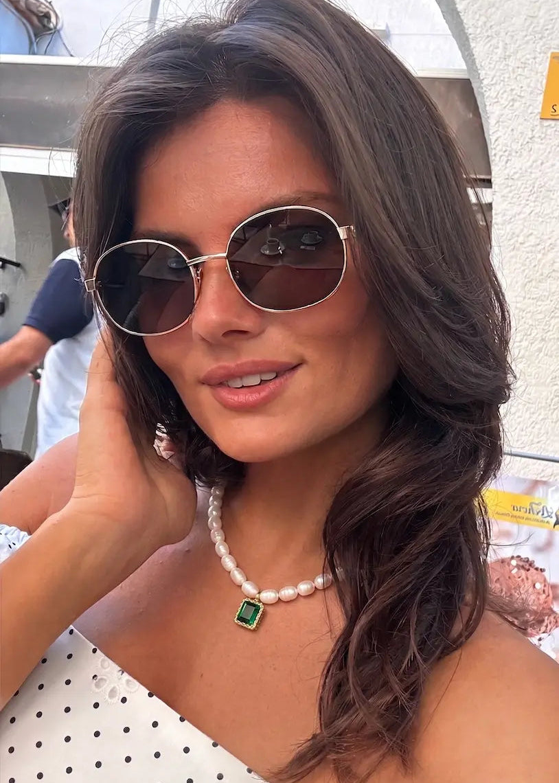 A woman wearing oval sunglasses with gold-toned metal frames and dark brown lenses, along with a pearl necklace featuring a green square pendant, in an outdoor setting.