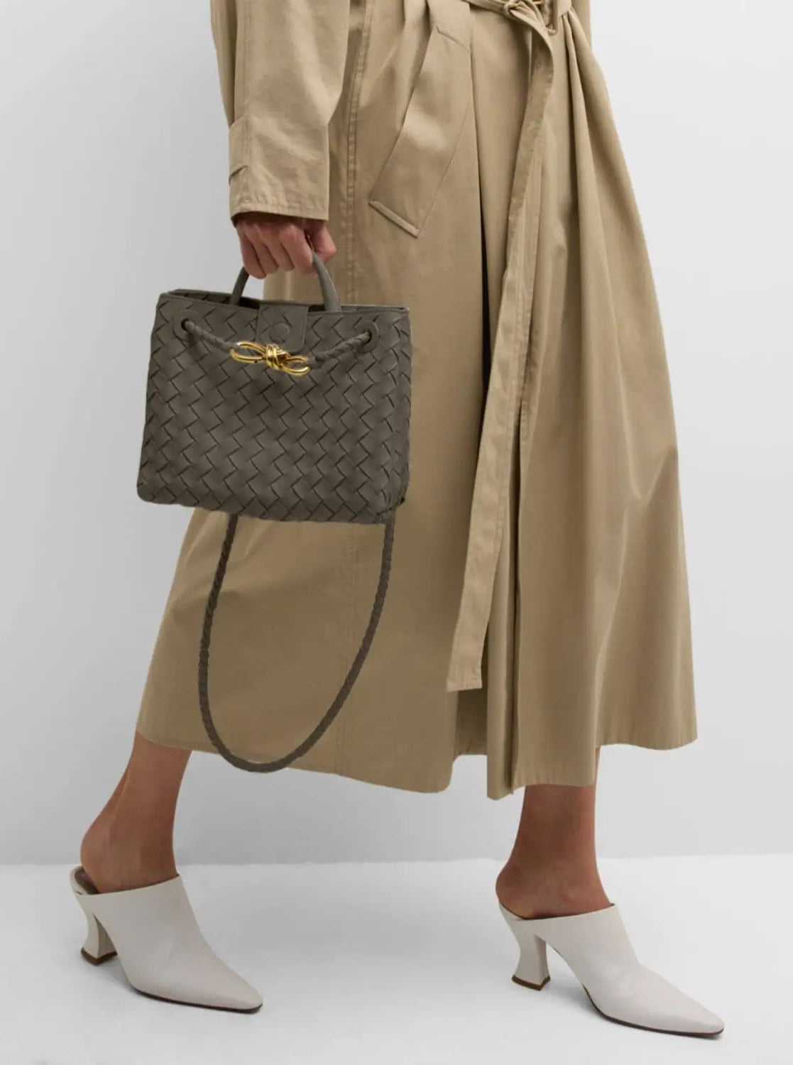 Person wearing a beige trench coat holding a woven handbag on a white background