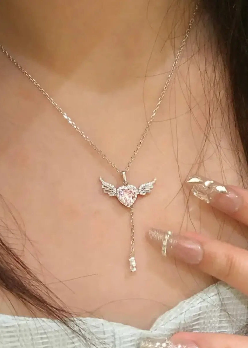 A close-up of a person wearing a delicate silver-toned necklace with a winged heart pendant. The pendant features a clear rhinestone-encrusted heart with small wings and a dangling clear rhinestone drop.