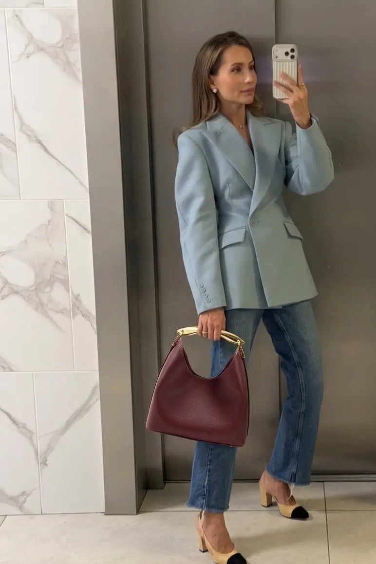 Woman taking a mirror selfie wearing a light blue blazer and jeans, holding a maroon handbag.