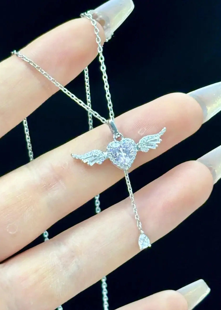 A close-up of a hand with long clear acrylic nails holding a silver-toned angel wing pendant necklace with a heart-shaped clear gemstone.