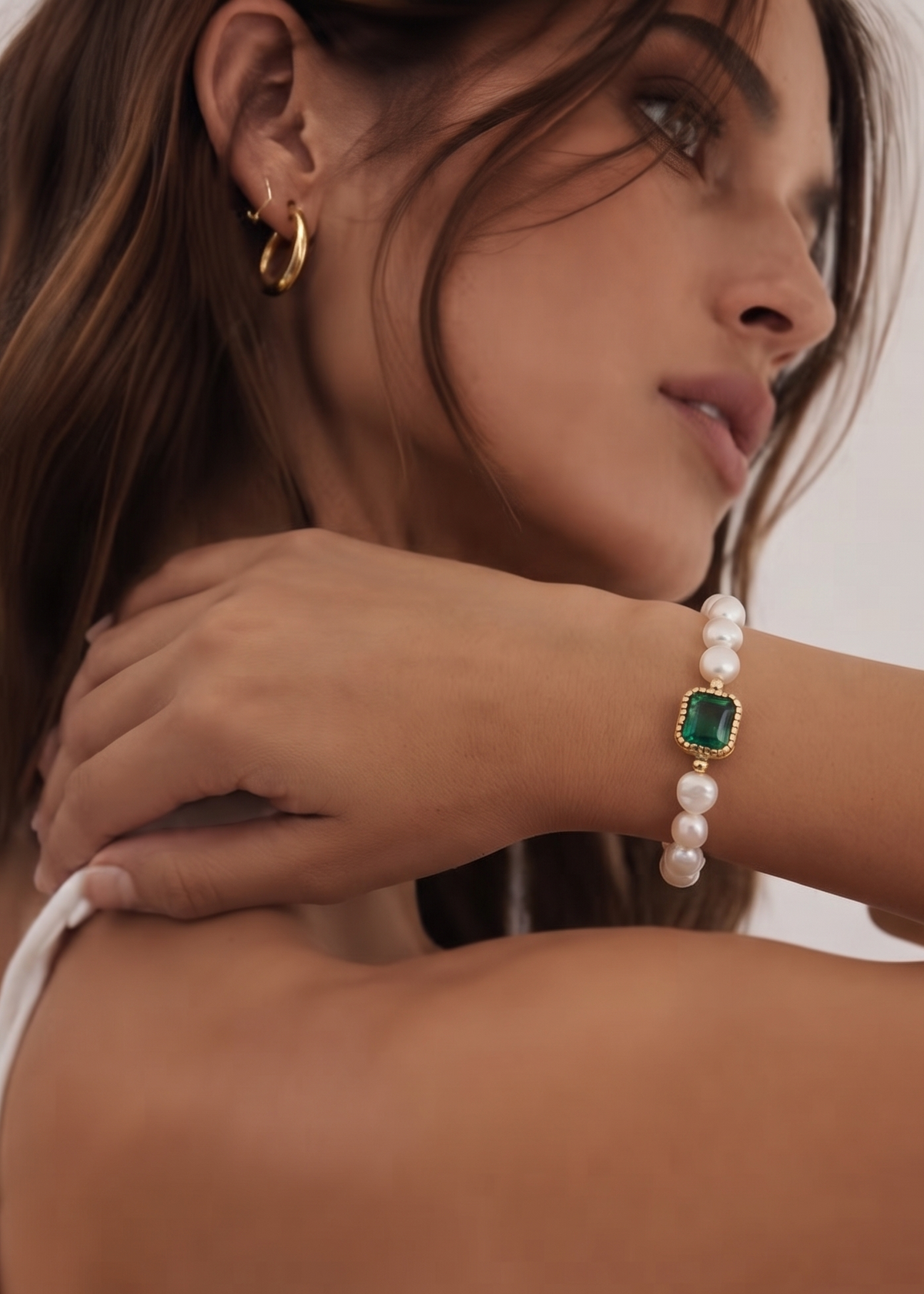A close-up lifestyle shot of a model wearing a gold-plated bracelet with green square gemstones and white pearls, paired with a gold huggie hoop earring.