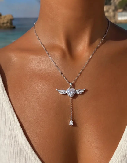 A silver angel wing pendant necklace with a heart-shaped center and a dangling teardrop charm, shown worn on a model's neck against a beach background.