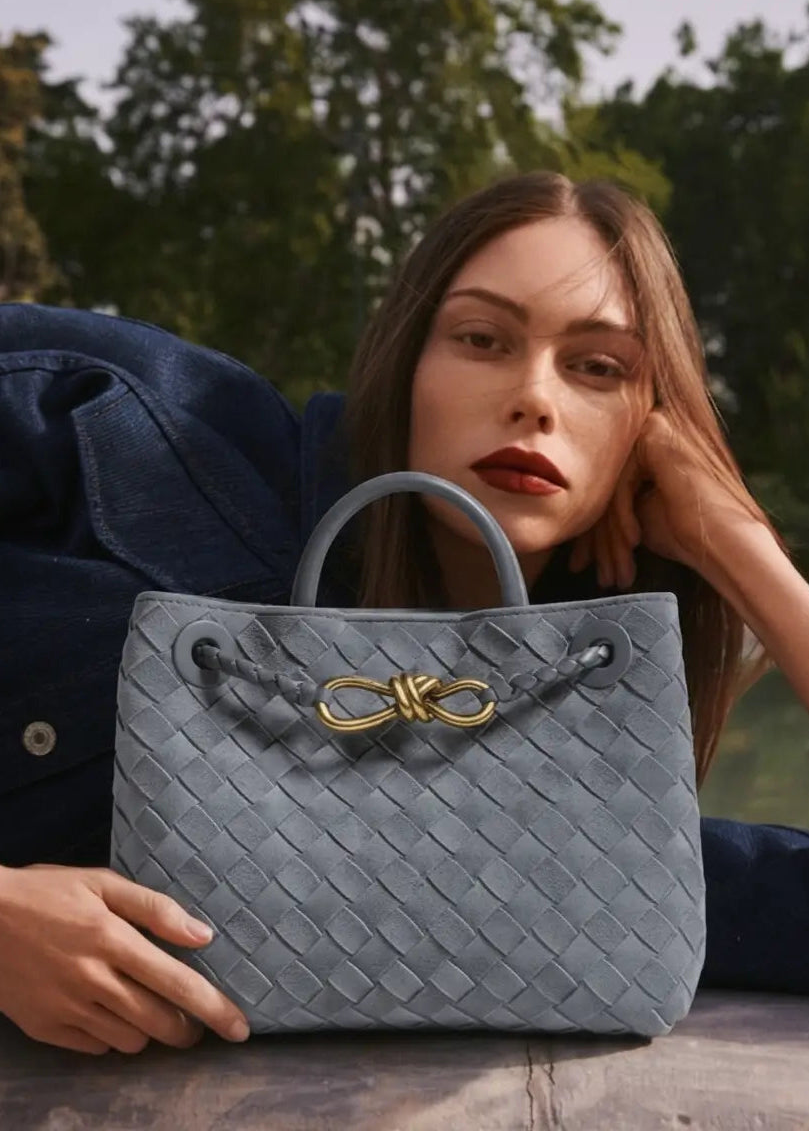 Woman holding a gray textured handbag with a gold bow detail outdoors.