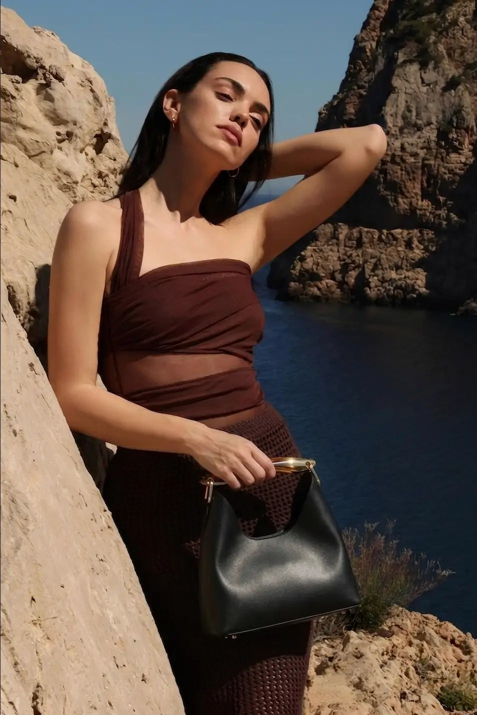 Woman in a brown dress holding a black handbag against a rocky cliff backdrop