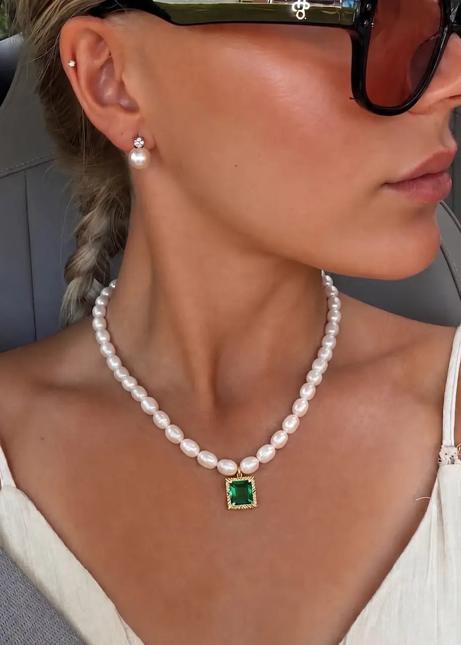 A close-up lifestyle shot of a woman wearing a white pearl necklace with a square green gemstone pendant in a gold setting, along with matching pearl stud earrings.