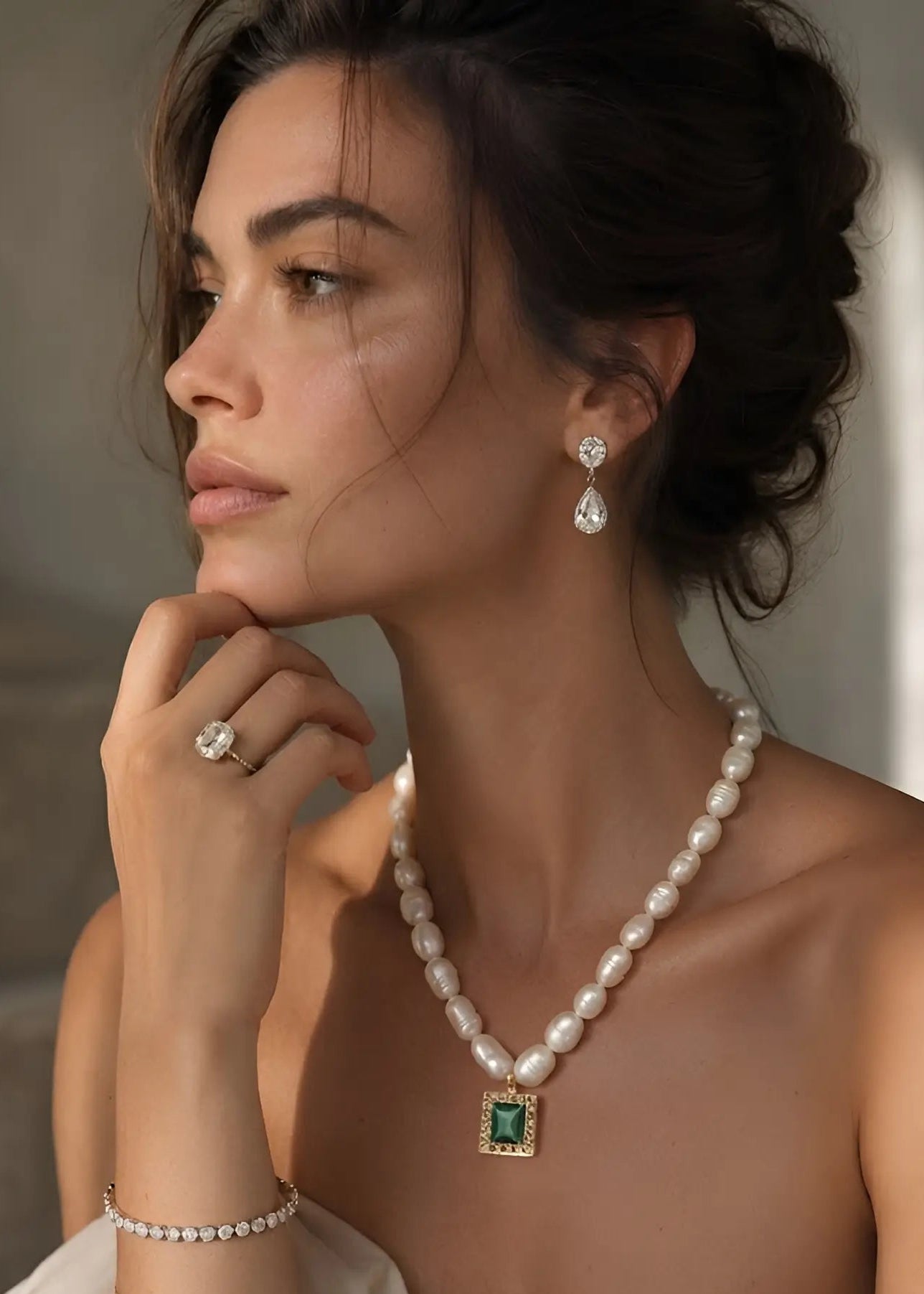 A woman in profile wearing a pearl necklace with a square green gemstone pendant, matching pearl drop earrings, and a pearl ring, styled with a cream draped garment against a soft neutral background.