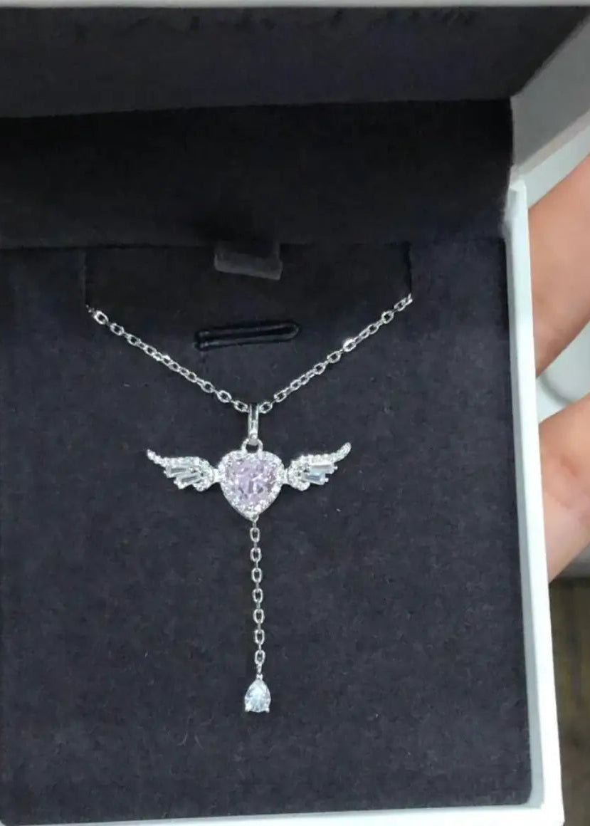 A silver-toned angel wing pendant necklace with a heart-shaped center and dangling charm, displayed inside a black jewelry box held by hands.