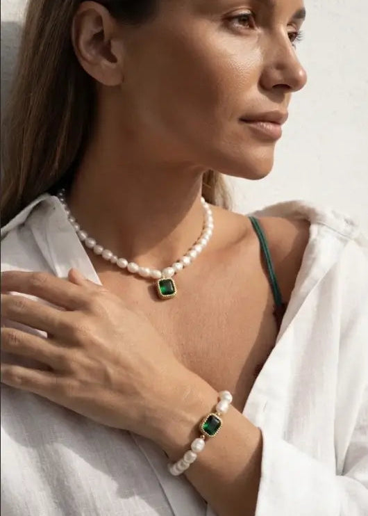 A woman wearing a white pearl necklace with a square green pendant and a matching white pearl bracelet with a green square accent, styled with a white shirt.