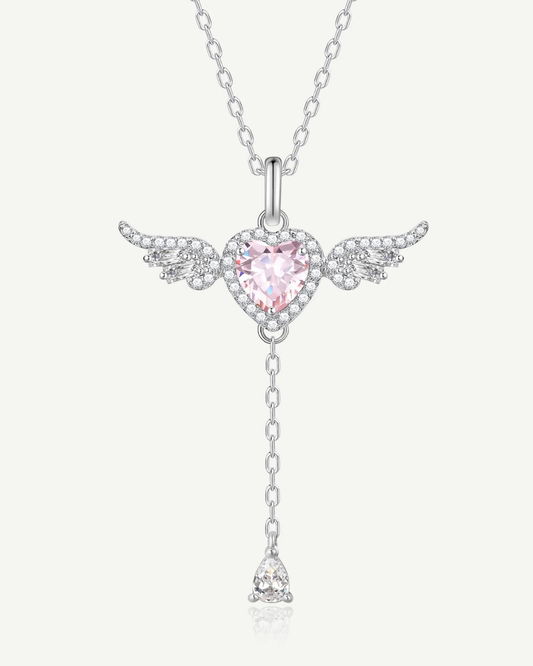 A silver-toned angel pendant necklace featuring a heart-shaped pink gemstone surrounded by small clear stones, with wings and a dangling chain ending in a teardrop-shaped clear stone, shown on a light off-white background.
