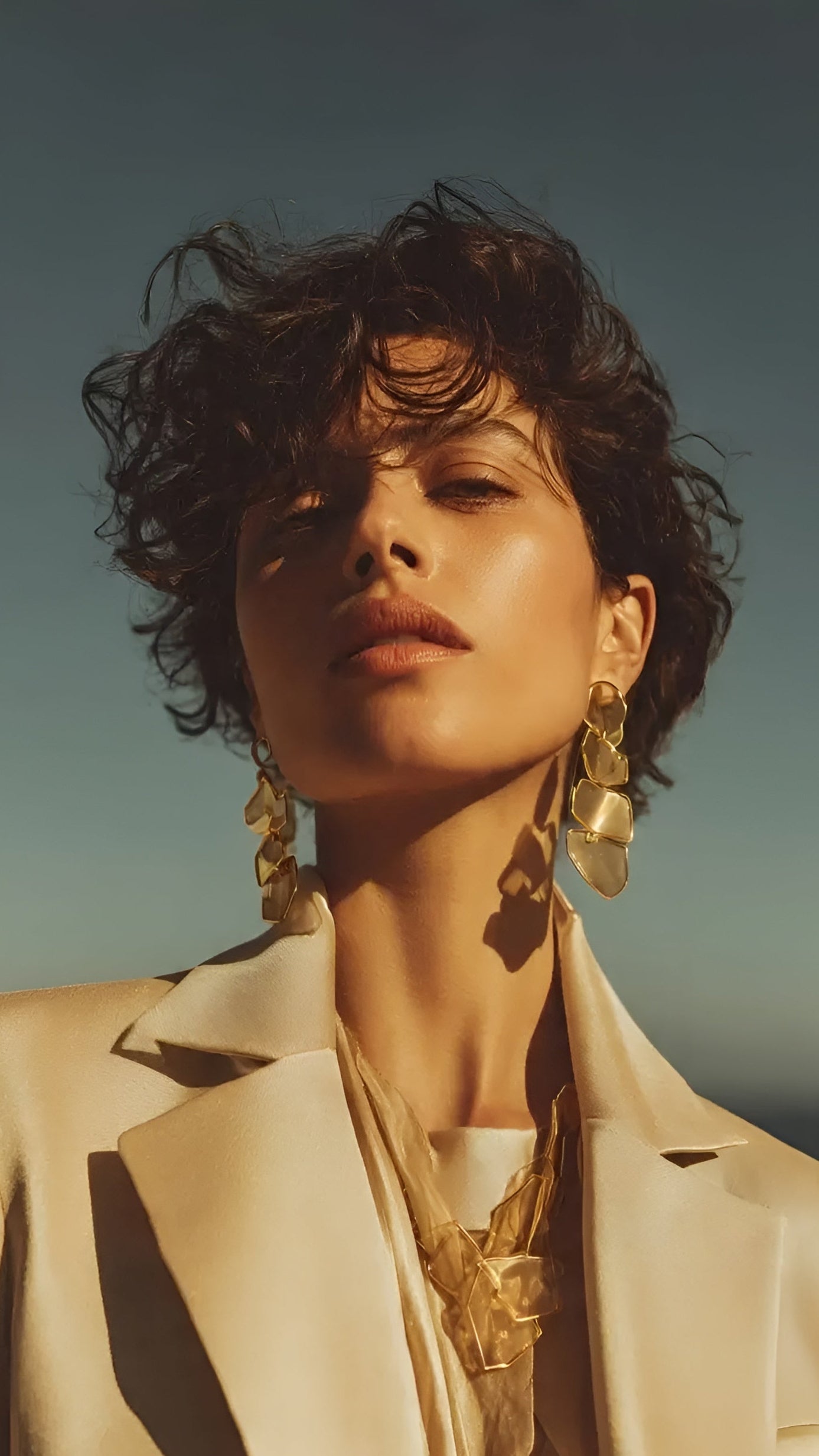 A close-up portrait of a woman with short curly dark hair, wearing large gold statement earrings and layered gold necklaces, dressed in a light beige blazer against a blue sky background.