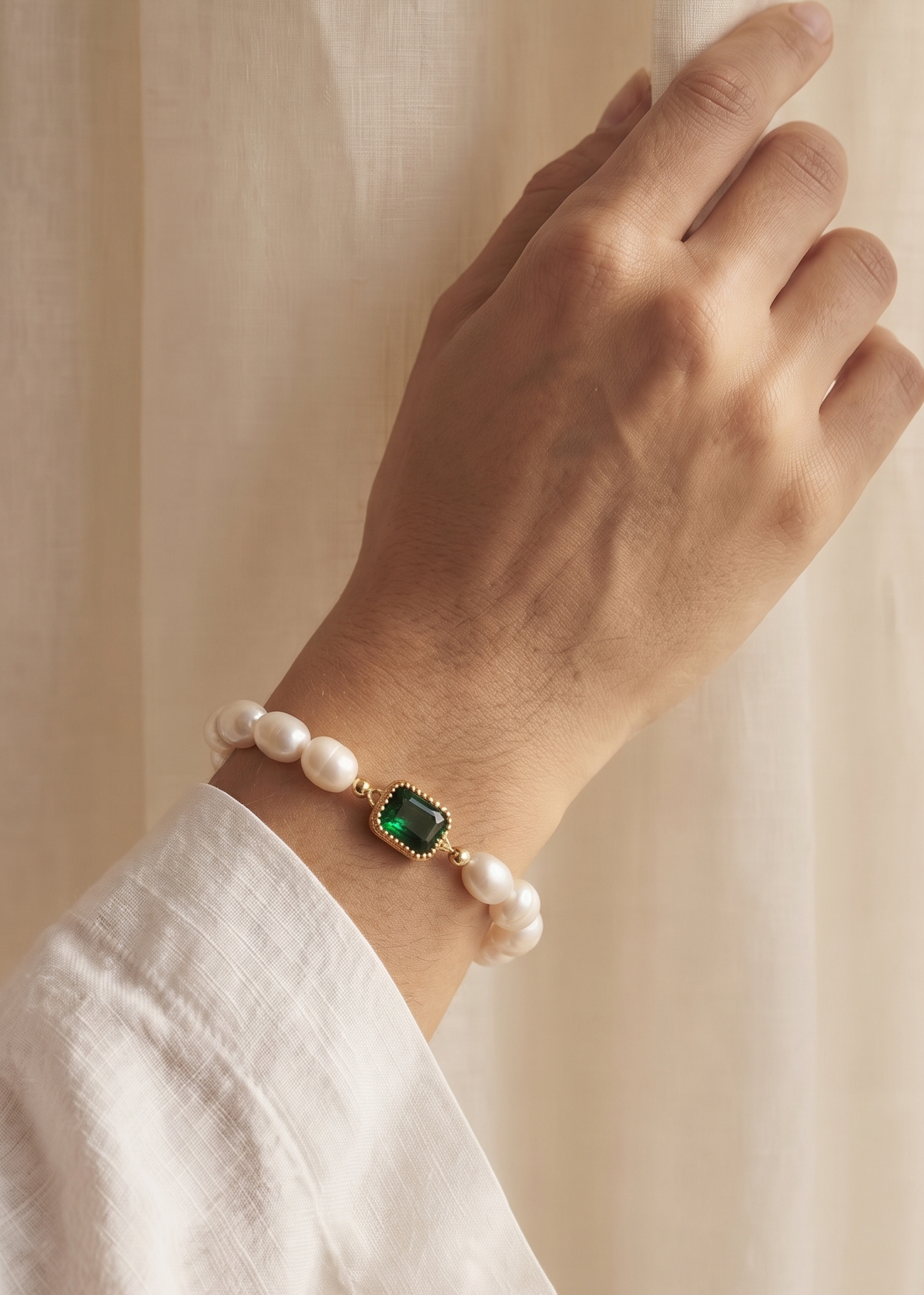 A close-up of a person's hand resting against a beige curtain, wearing a delicate bracelet made of white pearls and a central green rectangular stone set in gold.